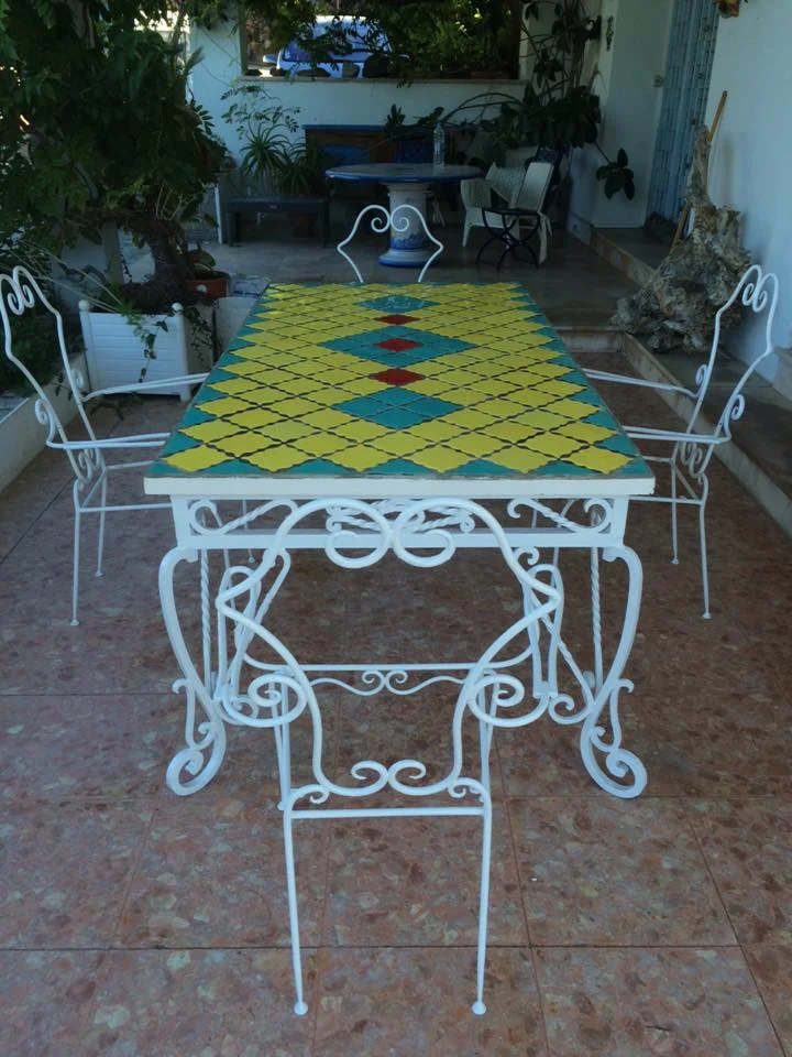 Table de jardin en fer forgé blanc avec plateau en carreaux de faïence à motifs colorés sur une terrasse carrelée.